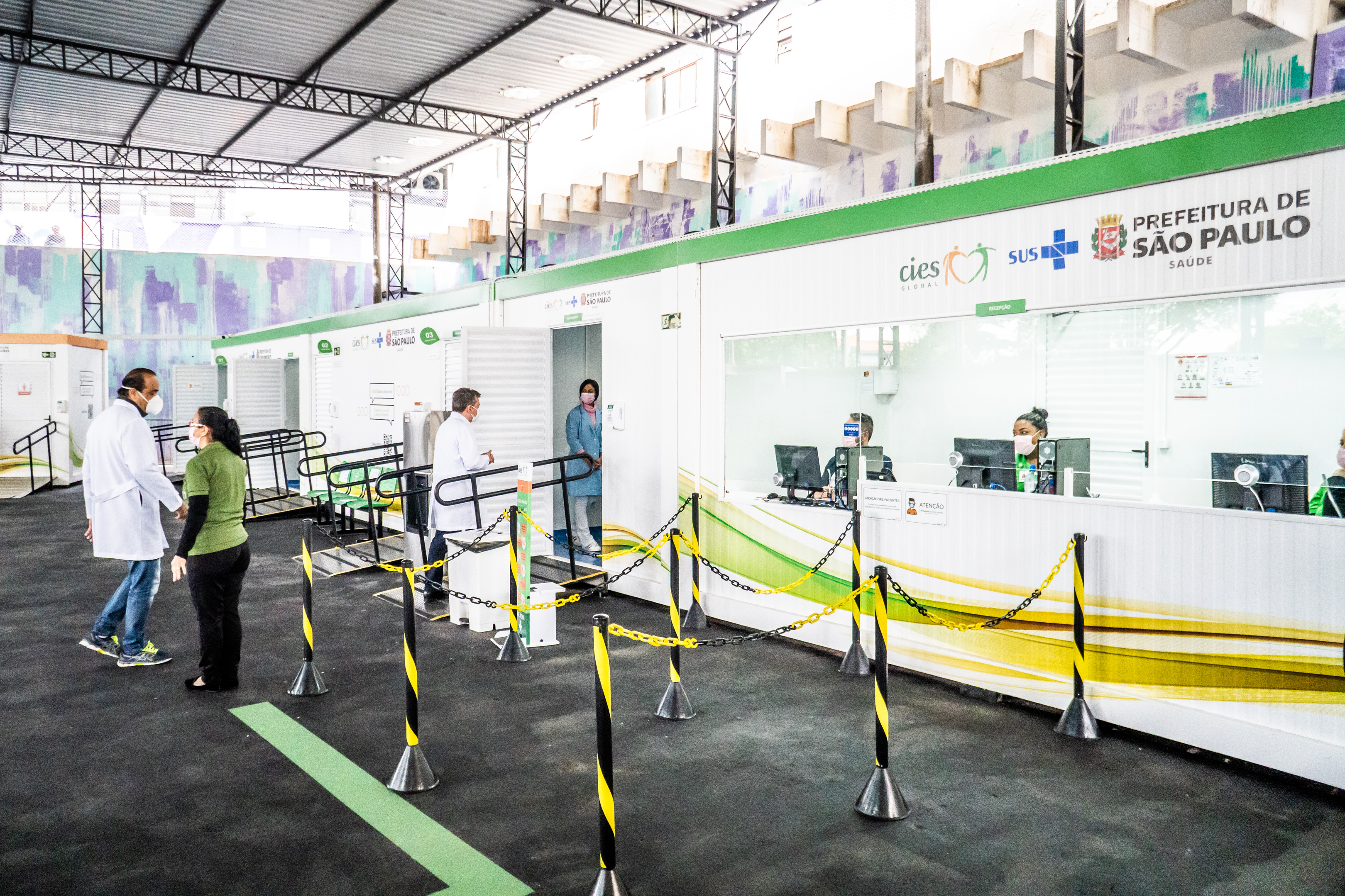 FOTO DE RECEPÇÃO -  Em primeiro plano temos uma longa mesa com recepcionistas  uniformizados, sentados diante do computador, ao fundo médicos de jaleco adentram às salas que se formam nos contêineres